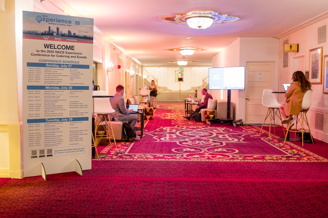 hallway with red carpet. Along walls of hallway are lounge chairs, short tables, lamps, and there are people looking at their laptops or phones while sitting on the furniture.