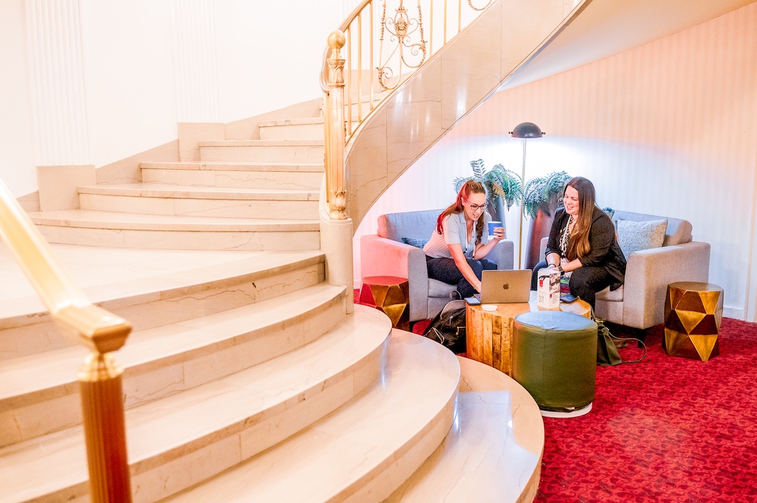 two women sit and talk with each other at lounge chairs in an area by a curved staircase. 