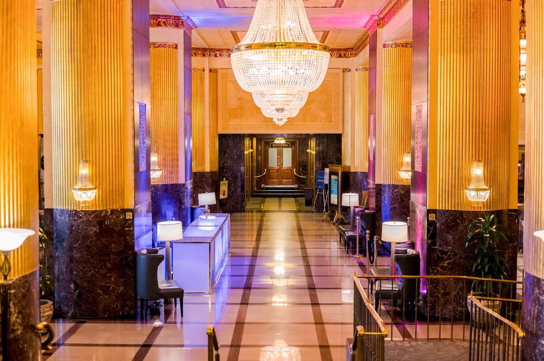 grand lit lobby of Milwaukee Hilton with tall columns and chandeliers. Columns lit in pink and blue. Light up registration desk sits in middle ground of lobby.