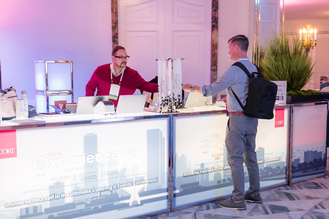 man in red sweater behind lit up branded registration desk hands something to a man in a blue shirt and gray pants on the other side of the registration desk