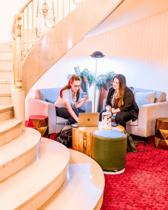 Two women sit in a lounge chair setup at base of curved staircase